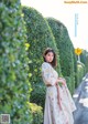 A woman in a white dress standing in front of a hedge.